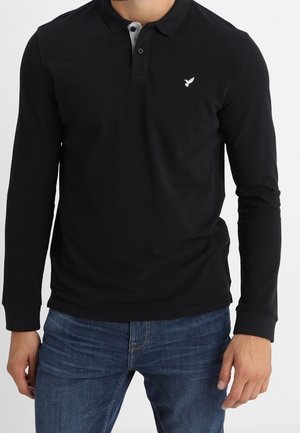 Polo noir à manches longues avec col boutonné et intérieur gris. Présente un petit logo blanc sur la poitrine gauche. Associé avec un jean bleu.