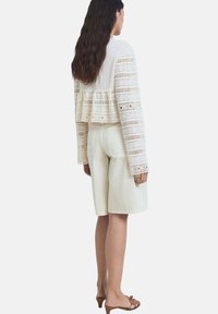 Giacca cropped a maglia all'uncinetto color crema con maniche lunghe, abbinata a pantaloni corti beige chiaro dal taglio sartoriale, che mostrano un fit rilassato e una texture liscia.