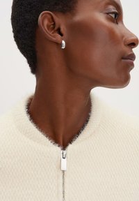 Witte getextureerde jack met ritssluiting aan de voorkant en een ronde kraag, gedragen met een zilveren oorbel. Close-up van het profiel van het model, met nadruk op detail en afwerking.