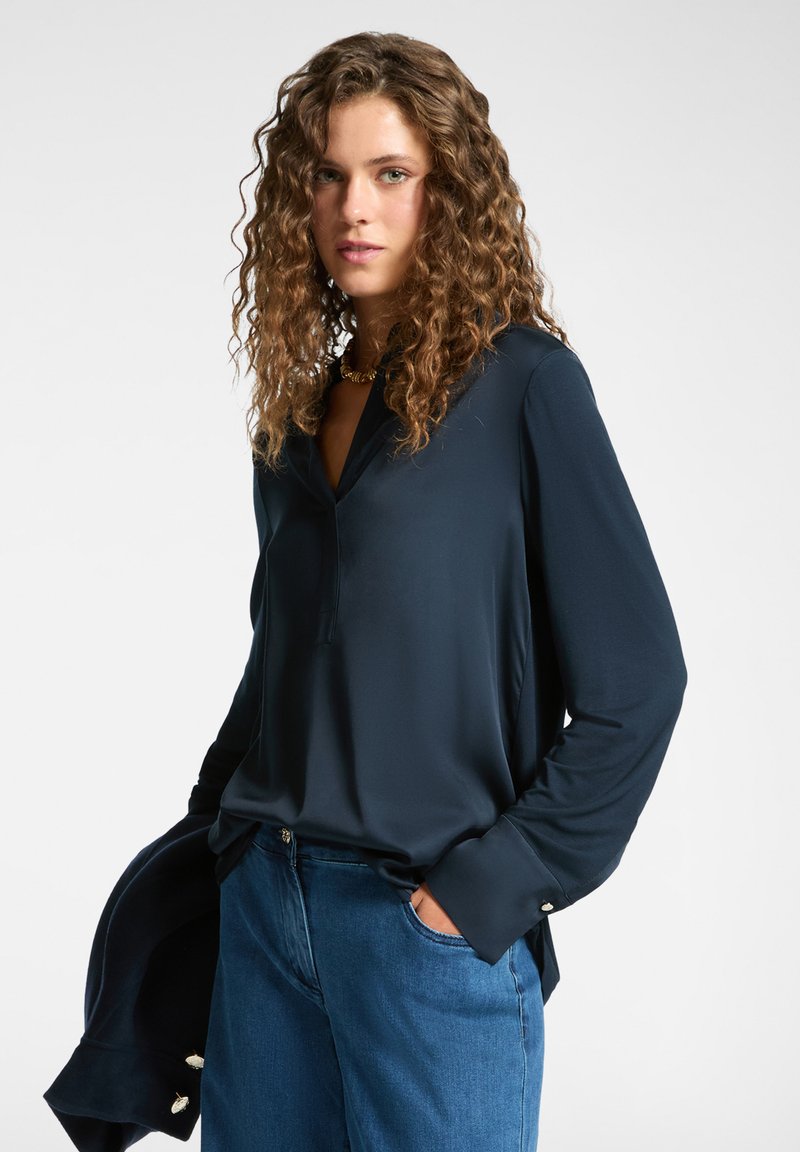 Tummansininen pitkähihaisen pusero, jossa on V-pääntie ja napitettavat hihansuut, valmistettu sileästä kankaasta, yhdistettynä sinisiin farkkuihin.