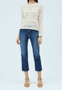 Krämfärgad stickad tröja med öppen väv, kombinerad med avkortade blå denimjeans. Modellen bär tanfärgade klacksandaler. Enkelt, avslappnat outfit.