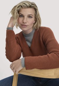 Ein rostfarbener, gestrickter Pullover mit V-Ausschnitt, der über einem grauen Hemd getragen wird. Das Model hat die Arme auf einen Holzstuhl gelegt, wodurch die strukturierten Ärmel hervorgehoben werden.