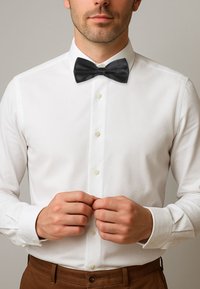 Witte overhemd met een zwarte satijnen strik. Het overhemd heeft knopen aan de voorkant en details aan de manchetten, gecombineerd met bruine broek.