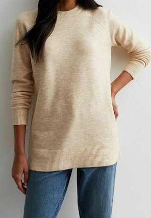 Femme portant un pull en tricot beige et un jean bleu, debout avec une main sur la hanche et l’autre relâchée sur le côté.