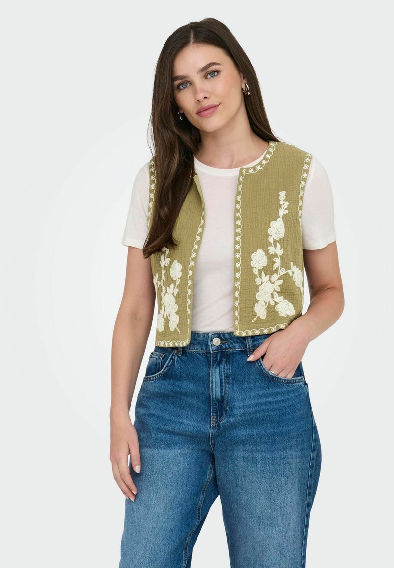 Olivgrüne, verkürzte Weste mit weißer Blumenstickerei und gewellten Kanten. Getragen über einem weißen Hemd und kombiniert mit blauen, hoch taillierten Jeans.