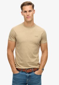 Beige t-shirt met korte mouwen en een ronde halslijn, met een subtiele logo op de linker borst. Draagt zich met blauwe spijkerbroek en een brown riem.