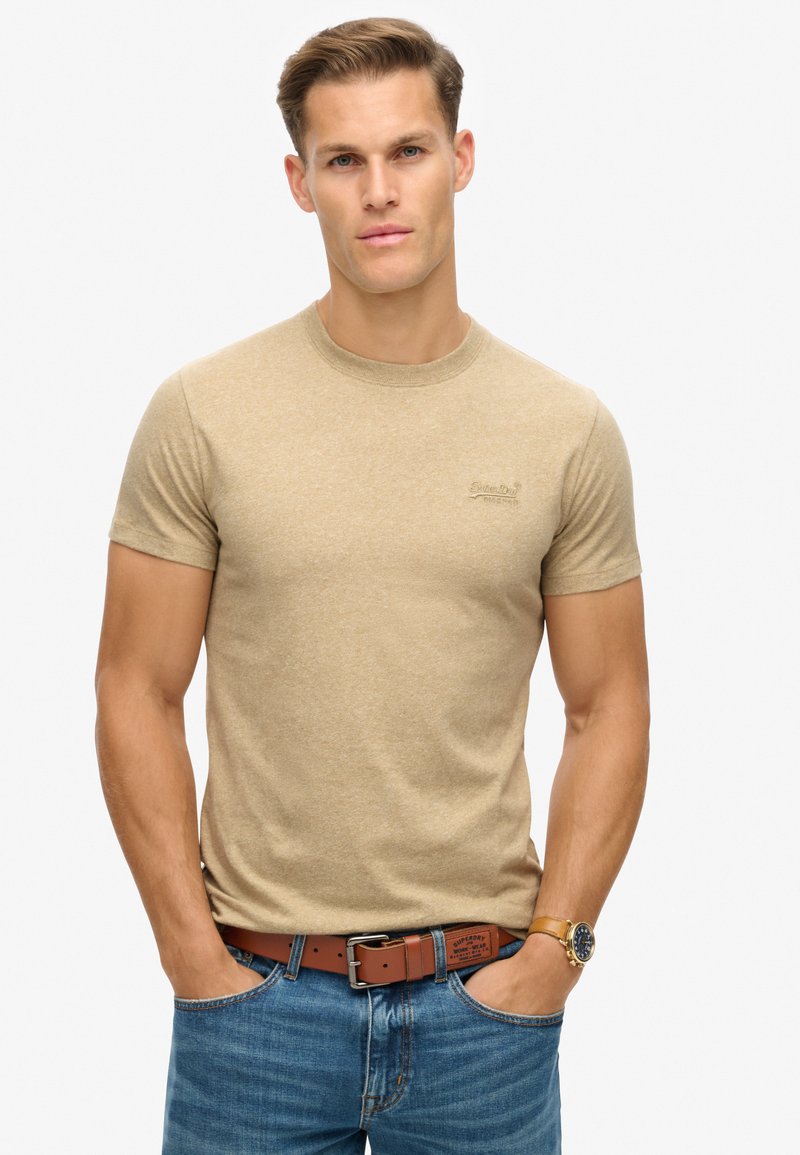 Beige t-shirt met korte mouwen en een ronde halslijn, met een subtiele logo op de linker borst. Draagt zich met blauwe spijkerbroek en een brown riem.