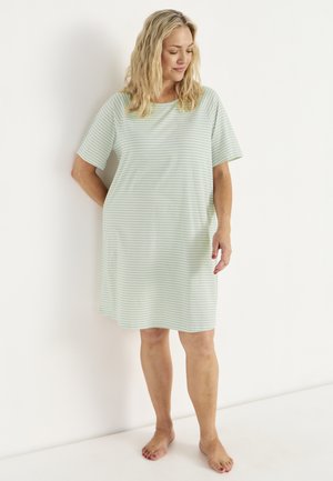 Vrouw staat blootsvoets in een losse, kortere mouw, knielange jurk met lichtgroene en witte strepen tegen een effen witte achtergrond.