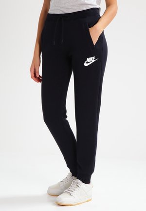 Personne portant un pantalon de survêtement noir Nike avec un logo blanc, debout avec une main dans la poche et des baskets blanches sur un fond clair.