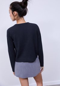 Pull en maille noire à manches longues et coupe courte, porté sur une mini-jupe grise texturée. Fond neutre mettant en valeur la tenue.