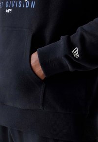 Hoodie nera realizzata in tessuto morbido, con tasca centrale, polsini a coste e accento del logo sul polsino. Stampa di testo sopra in blu scuro.
