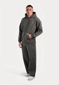 Grauer Kapuzenpullover mit Reißverschluss und Fronttaschen sowie passende graue Jogginghose. Der Stoff wirkt weich, mit dezenten strukturierten Akzenten. Schlichte weiße Turnschuhe.