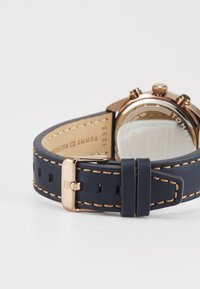 Montre avec un bracelet en cuir bleu marine mettant en valeur des surpiqûres orange et une boucle en or rose, montrée de dos sur une surface blanche.