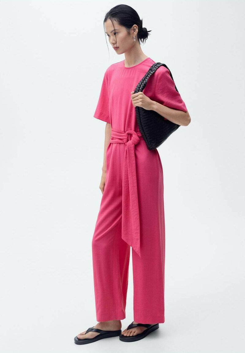 Rosa jumpsuit med korte ærmer, bæltet talje, lige ben, parret med en struktureret sort taske og sorte flip-flops. Glat stoffer.