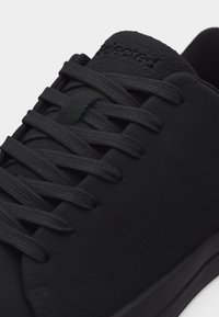 Gros plan sur une basket noire mettant en avant les lacets texturés et le logo en relief sur la languette.