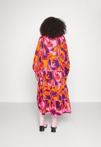 Een lange, kleurrijke jurk met een roze, oranje en paarse bloemenpatroon, met een gelaagd ontwerp en lange mouwen, gecombineerd met roze hoge laarzen tot de knie.