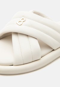 Κοντινό πλάνο λευκού σαγιονάρου με επένδυση και σταυρωτούς ιμάντες με χρυσό γράμμα «B» στον πάνω ιμάντα.