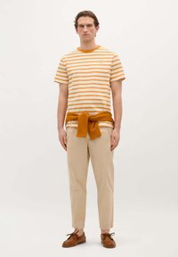 Camiseta de rayas en blanco y naranja, combinada con pantalones beige. Suéter de punto naranja atado a la cintura. Zapatos marrones con cordones.