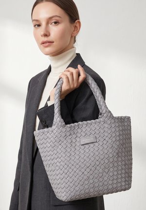 Femme en blazer sombre et col roulé blanc tenant un grand sac à main gris tressé avec des anses courtes et une étiquette de marque.