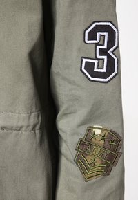 Manga de una chaqueta de estilo militar verde con un parche negro y blanco del número 3 y un parche de insignia "ARMY" en camuflaje que presenta estrellas y chevrones.