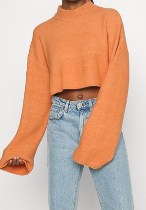 Avskuren orange tröja med långa, vida ärmar och hög krage, tillverkad av ett mjukt, texturerat material, kombinerad med höga ljusblå jeans.