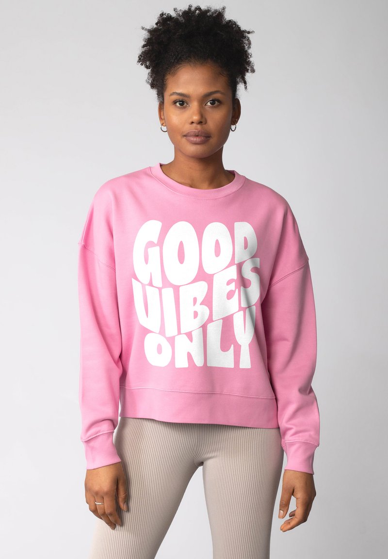 Vrouw met krullend haar draagt een roze sweatshirt met de tekst "GOOD VIBES ONLY" in het wit, gecombineerd met beige geribde leggings, staande tegen een neutrale achtergrond.