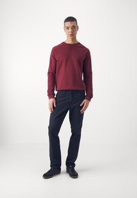 Hanorac maron cu guler rotund și manșete ribbate, asociat cu blugi închiși la culoare și adidași negri. Modelul stă în fața unui fundal alb simplu.