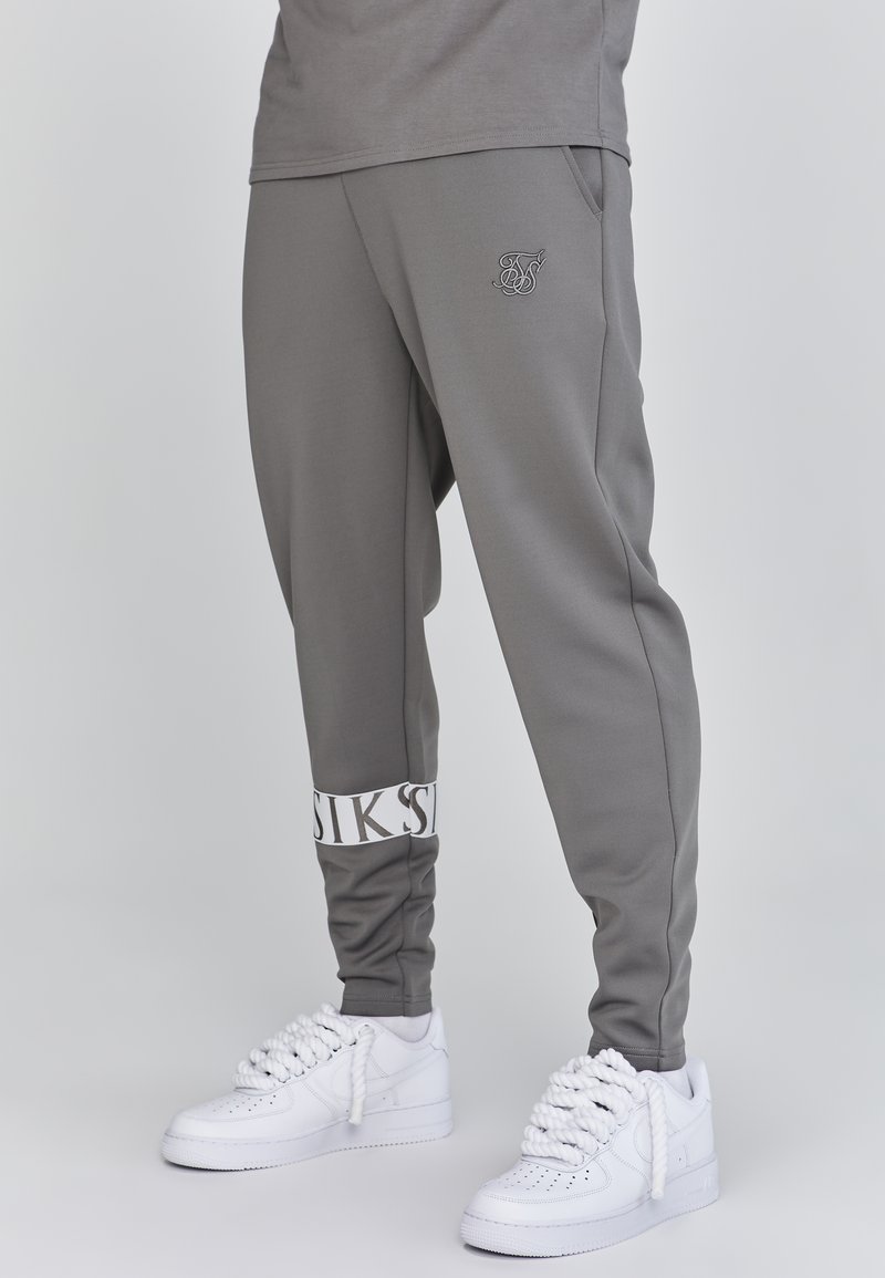 Grijze sportbroeken in een taps toelopend ontwerp met zijzakken, voorzien van een witte gebrandmerkte boord en logo op het bovenbeen, gecombineerd met witte sneakers.