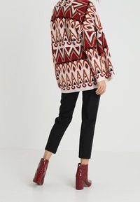 Stickad kofta med röda, svarta och krämfärgade geometriska mönster; kombinerad med svarta smala byxor och texturerade bordeauxfärgade ankelskor.