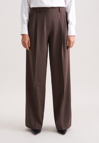Bruine hoge taille broek met een wijd uitlopende snit, voorzien van plooien en een elegante textuur. Gecombineerd met puntige zwarte schoenen.