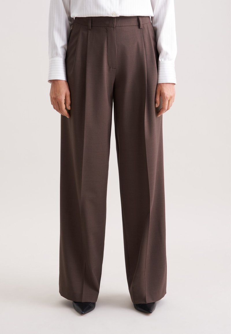 Bruine hoge taille broek met een wijd uitlopende snit, voorzien van plooien en een elegante textuur. Gecombineerd met puntige zwarte schoenen.