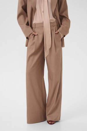 Donna che indossa pantaloni a gamba larga color beige e blazer abbinato con le mani nelle tasche, abbinati a tacchi aperti color burgundy.