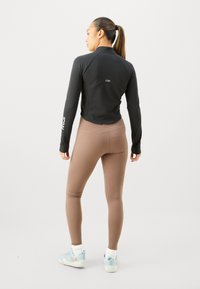Kvinna med hästsvans som bär en svart långärmad topp, bruna leggings, vita strumpor och ljusblå sneakers, stående med ryggen mot kameran på en enfärgad bakgrund.