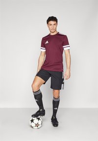 Bordeaux sportshirt met witte gestreepte mouwen, gecombineerd met zwarte shorts en kniehoge zwarte sokken. Zwarte voetbalschoenen en bal inbegrepen.