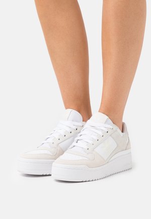Een paar benen met witte Adidas-sneakers met suède- en lederen details, dikke zolen en veters gestrikt, tegen een effen witte achtergrond.