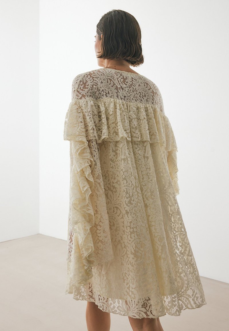 Kreemi pitseekleid, millel on lainelised varrukad, keerukad lillemustrid, põlvekohale ulatuv lõige ja voolav siluett. Ükslane taust.