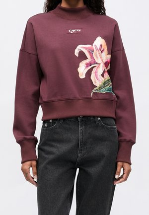 Femme portant un sweat-shirt Guess bordeaux avec un grand motif floral rose et blanc sur le devant, associé à un jean noir taille haute.