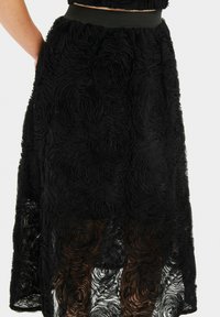 Fustă neagră de lungime până la genunchi, cu un model texturat în formă de trandafiri, dintr-un material semi-transparent. Talie elastică. Designul fluid accentuează mișcarea.