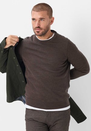 Mann mit kurzem Haar und Bart zieht eine dunkelgrüne, geknöpfte Jacke über einem braunen Pullover und einem weißen Unterhemd vor einem einfarbigen Hintergrund an.