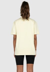 Light yellow cotton t-shirt with short sleeves and a relaxed fit, viewed from the back. Paired with black fitted shorts.