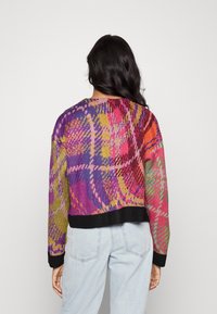 Kobieta z długimi ciemnymi włosami, ubrana w sweter z abstrakcyjnym wzorem w wielu kolorach, z czarnymi mankietami i jasnoniebieskimi dżinsami, stoi na jednolitym tle.