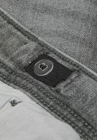 Tejido de mezclilla gris con una etiqueta de tela negra visible y un botón de plástico transparente. Presenta costuras cosidas y una superficie texturizada.