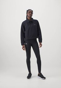 Zwarte cropped pullover met een kwart-ritssluiting, gecombineerd met zwarte legging. Model draagt zwarte sportschoenen. Gladde textuur, minimale branding.