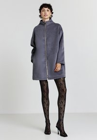 Donna in piedi che indossa un cappotto grigio con zip, collant neri a rete e sandali neri con tacco alto su uno sfondo chiaro e uniforme.