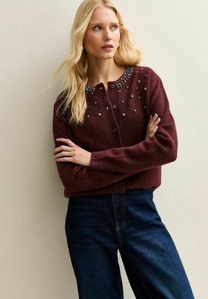 Cardigan en laine bordeaux avec col rond, fermeture à boutons et gemmes décoratives sur les épaules ; associé à un jean en denim foncé.