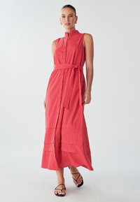 Červené maxi šaty bez rukávov s vysokým golierom, zapínaním na gombíky a opaskom s viazaním v páse. Majú tiered lem a mäkkú, hladkú látku.