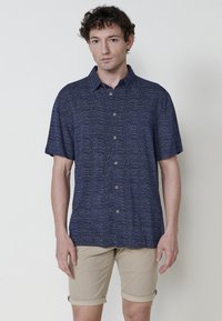 Camicia azzurra a maniche corte con un motivo testurizzato, apertura con bottoni e colletto classico, abbinata a pantaloncini chiari beige arrotolati.