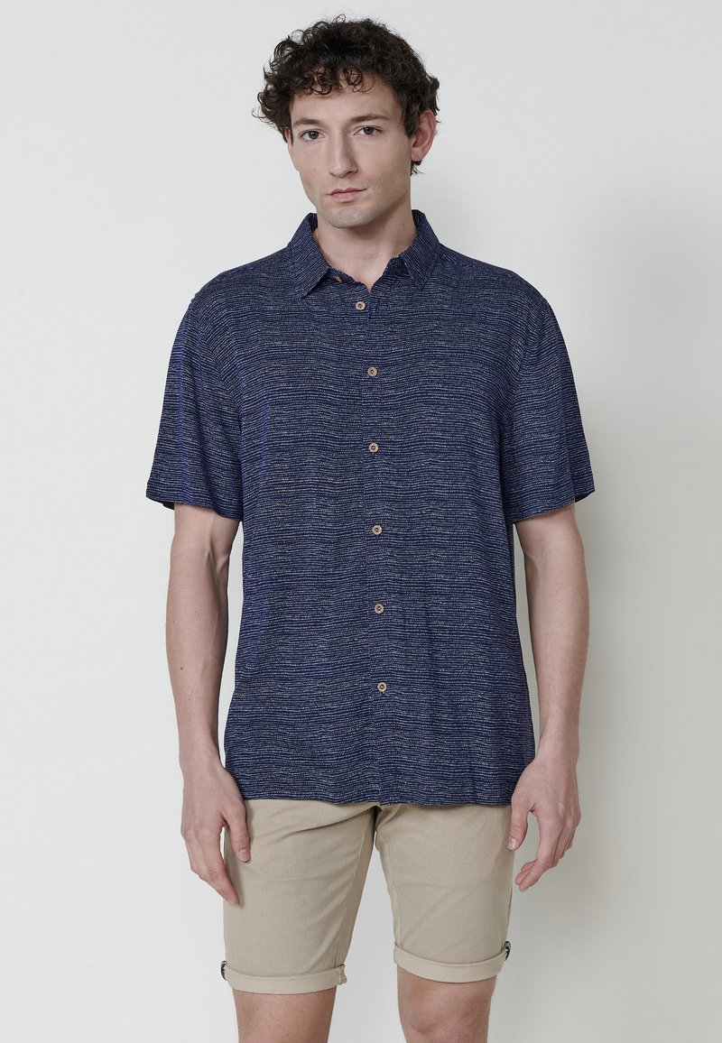 Camicia azzurra a maniche corte con un motivo testurizzato, apertura con bottoni e colletto classico, abbinata a pantaloncini chiari beige arrotolati.