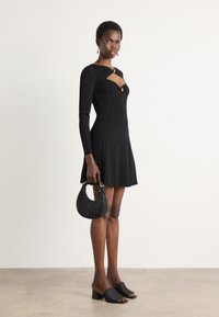 Vestido preto em malha com mangas longas, recorte em forma de olho de fechadura e detalhes em dourado. Combinado com uma mala preta texturizada e mules de salto. Fundo simples.