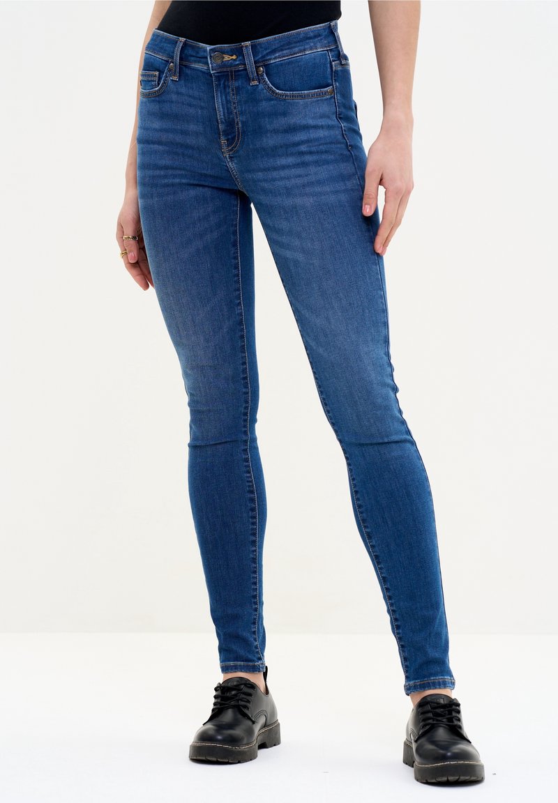 Jeans ajustados azul oscuro de denim, con bolsillos frontales, cierre con botón y un ligero desvanecido, combinados con zapatos negros de cordones.